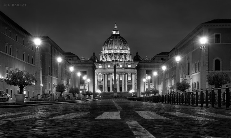 Wide, empty avenue leading up to St Peter's Basilica in the Vatican. The buildings are floodlit.