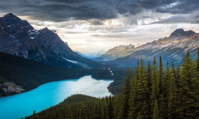 Large turquoise lake sits at the bottom of forested valley surrounded by majestic mountains lit by evening sun.