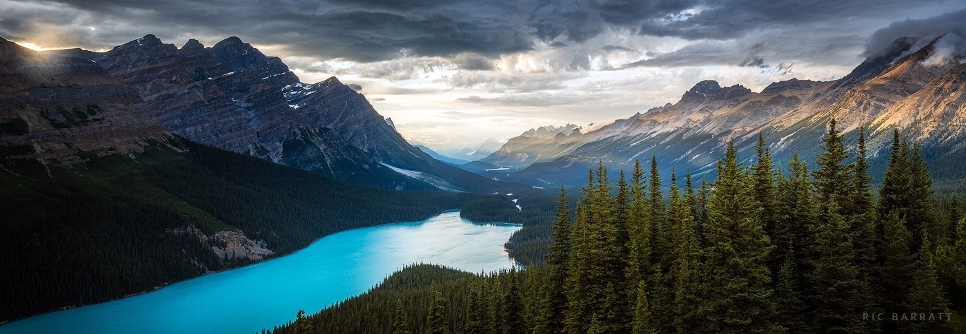 Large turquoise lake sits at the bottom of forested valley surrounded by majestic mountains lit by evening sun.