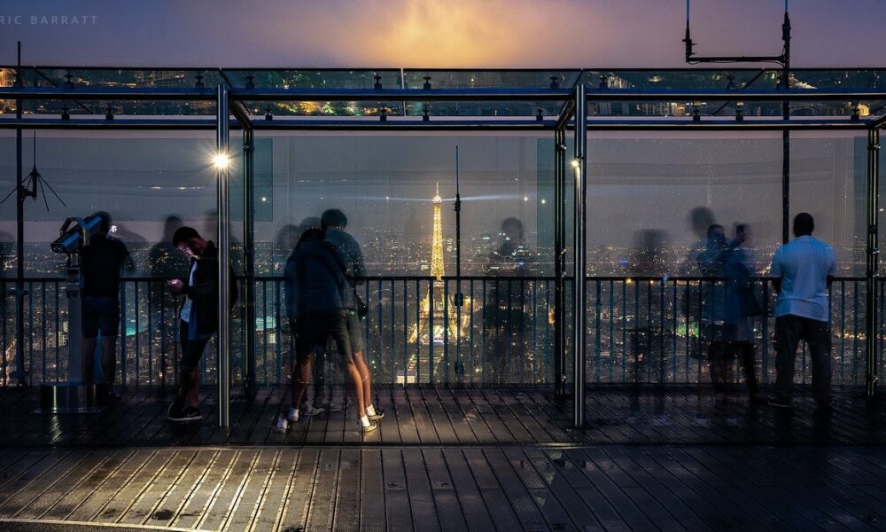 Tourists look out from a high viewing platform in Paris at night. Illuminated Eiffel Tower in the distance.