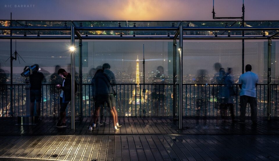 Tourists look out from a high viewing platform in Paris at night. Illuminated Eiffel Tower in the distance.