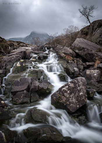 Small waterfall stream cascades down grey rocks in overcast mountainous terrain.