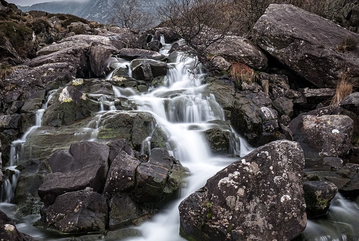 Small waterfall stream cascades down grey rocks in overcast mountainous terrain.