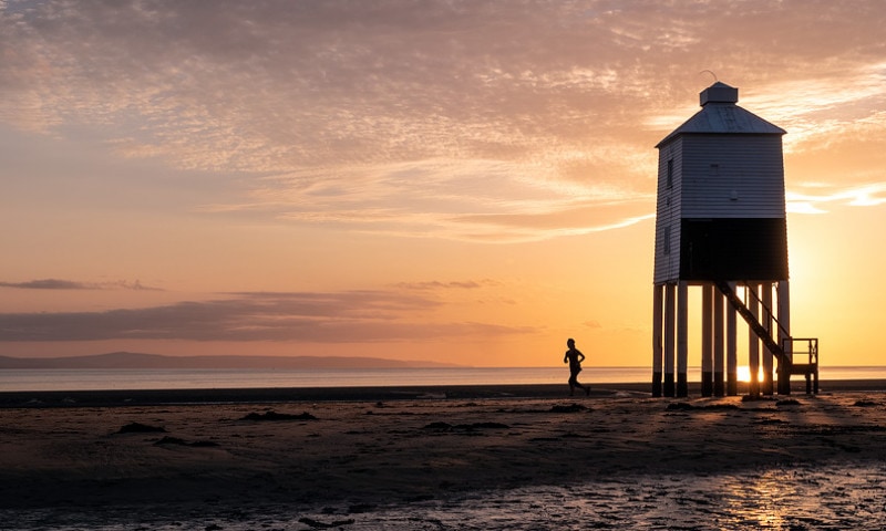 Runner runs past a stilted, wooden lighthouse at sunset.