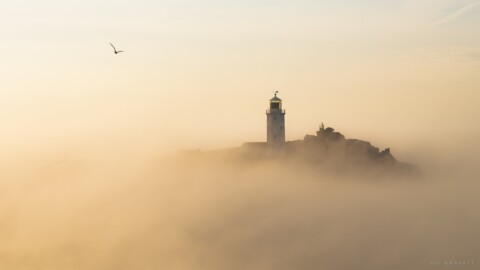 A lighthouse on a rocky island, partially obscured by clouds and sea mist.