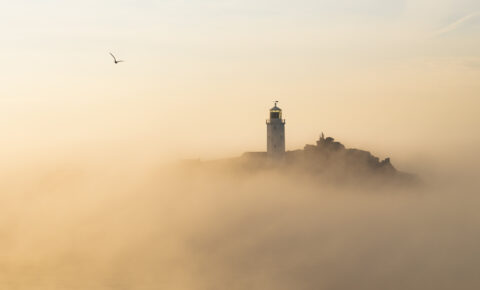 A lighthouse on a rocky island, partially obscured by clouds and sea mist.