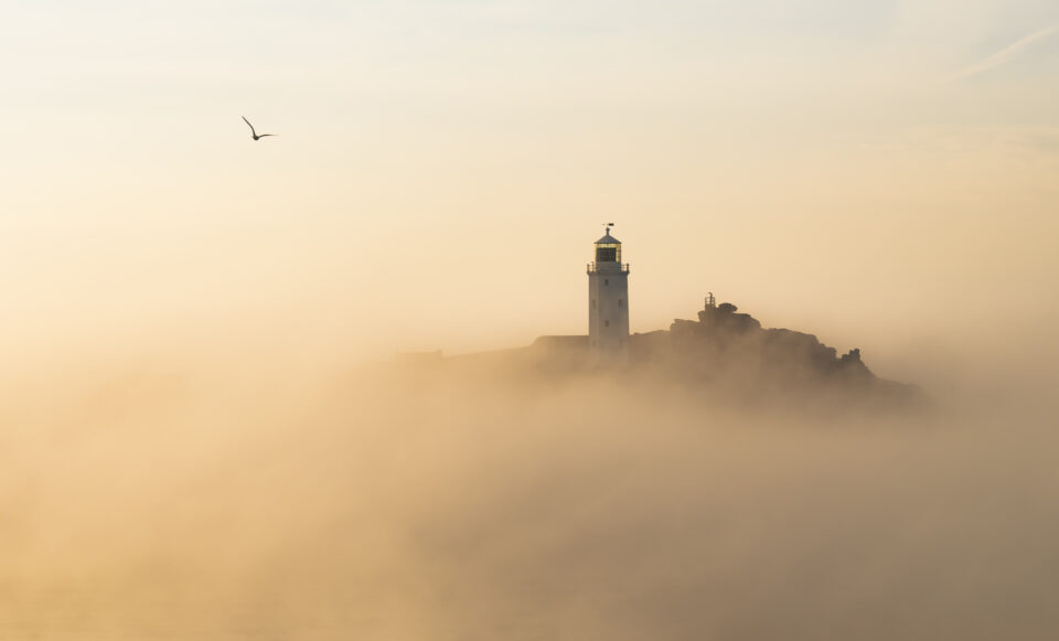 A lighthouse on a rocky island, partially obscured by clouds and sea mist.