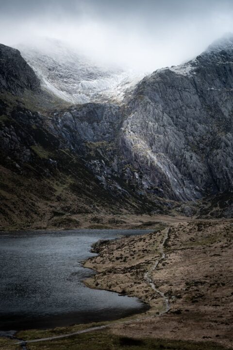 A lakeside path leads towards snowy mountain.