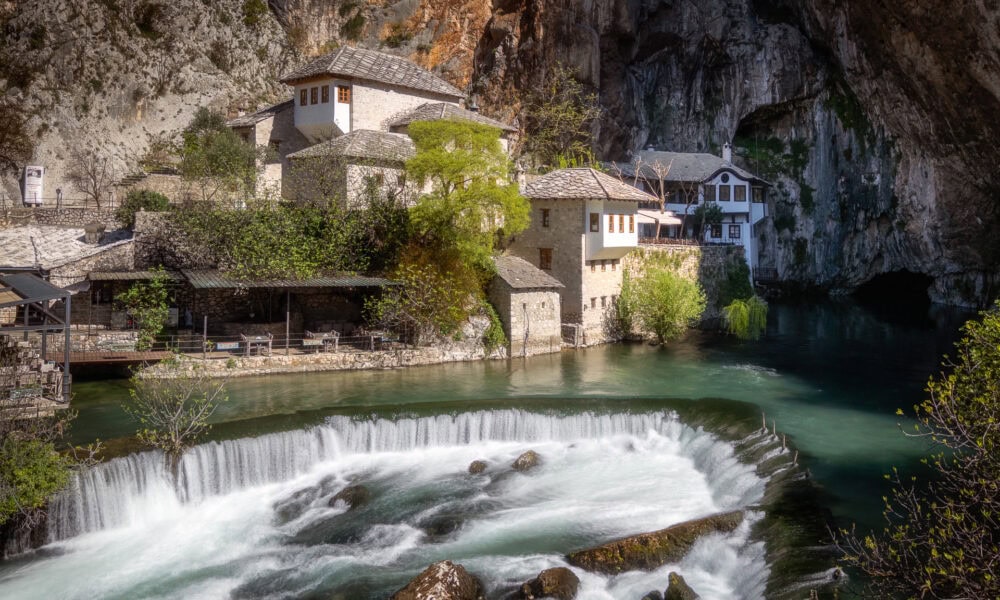 Serene view of Blagaj Tekija monastery nestled beside the river with cascading water and rocky cliff.