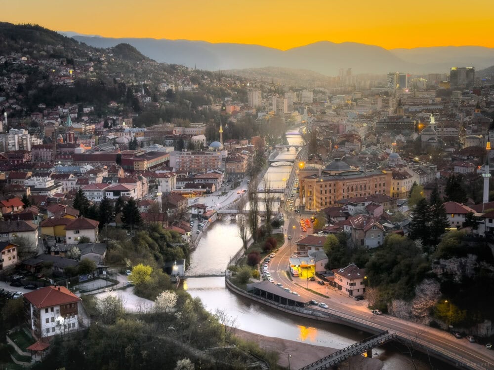 Sunset over Sarajevo with vibrant orange sky and cityscape illuminated at dusk.