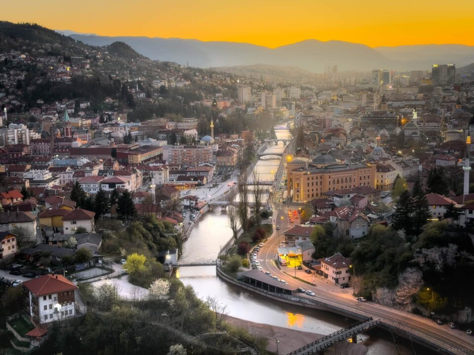 Sunset over Sarajevo with vibrant orange sky and cityscape illuminated at dusk.
