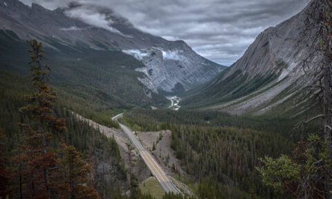Majestic mountain valley with winding road and dense forest under cloudy sky.