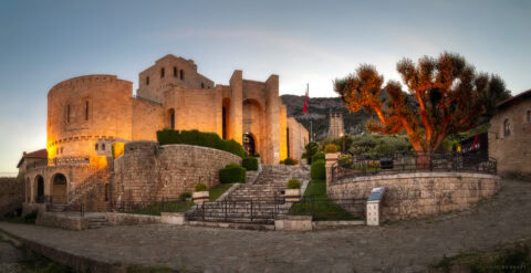 Castle fortress illuminated at sunset with warm lighting, stone walls, and surrounding greenery.