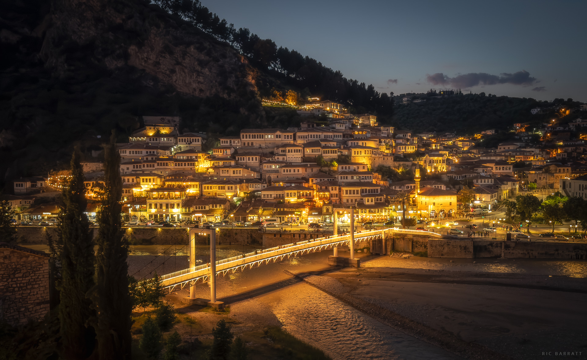 Night view of Berat with illuminated historic buildings and bridge over the river.