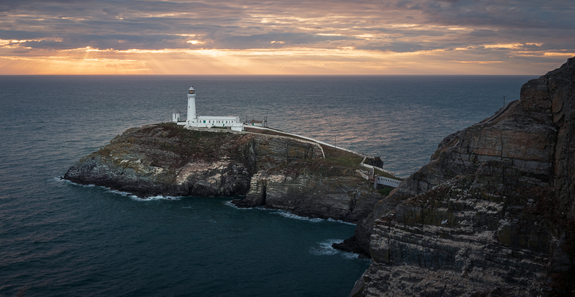 Lighthouse on a rocky island at sunset with dramatic sky and ocean.