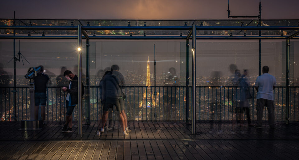 People viewing the Paris skyline from a rooftop observation deck at night.